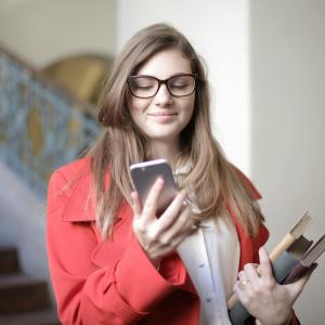 Pitkähiuksinen nuori henkilö katselee kännykkää. Toisessa kädessä on kirjoja. Taustalla julkisen tilan portaikko ja pylväs.