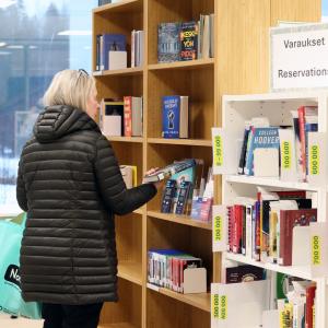 The customer is examining the back cover of a book and standing in front of the reservation shelf at Vuores Library.