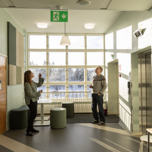 Kaksi henkilöä katsoo ylöspäin kohti kattovalaisimia koulun käytävän päässä ikkunan edessä.