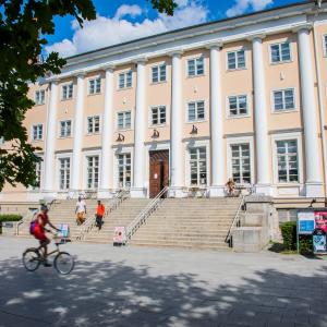 Wide steps lead up to a large light yellow building. People are walking in front of the building. The weather is sunny.