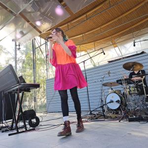 Pinkki-oranssiin mekkoon pukeutunut laulaja esiintyy ja mustiin pukeutunut rumpali soittaa taustalla.