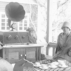 Mustavalkoisessa valokuvassa kaksi naista istuu ulkona kuuntelemassa vanhanaikaista radiota.