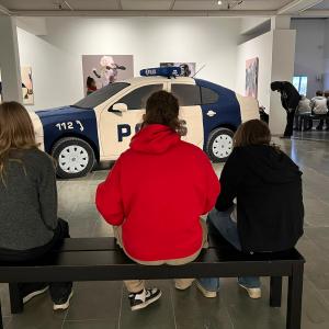 Kolme henkilöä istuu penkillä museossa. Taustalla teoksia ja poliisiautoteos.