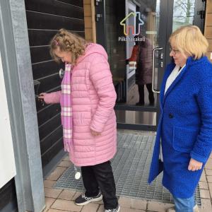 Vaaleanpunaiseen toppatakkiin pukeutunut kehitysvammainen nainen avaa avaimella kotioveaan. Hänen takanaan seisoo hänen äitinsä, jolla on sininen takki.