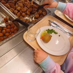 Pastellivärein liukuvärjättyyn huppariin pukeutunut lapsi on ottamassa lihapullia lautaselleen linjastossa.