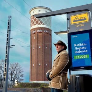 Cowboy-hattuinen mies nojaa pysäkkikatokseen, jossa näkyy mainos: Ratikkalinjasto laajenee 7.1.2025.