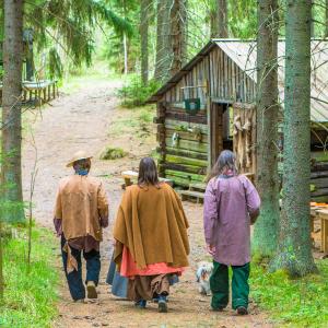 Kolme henkilöä kävelee metsätiellä selät kohti kameraa. Tien varralle on puisia rakennuksia.