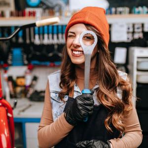 Pitkähiuksinen nuori henkilö kurkistaa toisella silmällä son kiintoavaimen reiän läpi. Taustalla pyöränkorjauspaja työkaluineen.