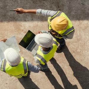 Kolme rakennusalalla työskentelevää henkilöä seisoo pihalla. Heidät on kuvattu ylhäältä päin. Päässä kypärät ja päällä huomioliivit. Yksi osoittaa kädellään eteenpäin, toisella on sylissä läppäri ja kolmannella papereita.