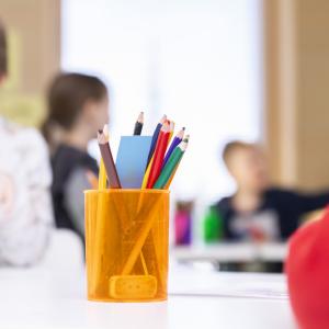 Kynäkuppi on pöydällä etualalla ja taustalla pieniä koululaisia.