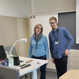 Two people are standing in a classroom by the teacher’s desk.