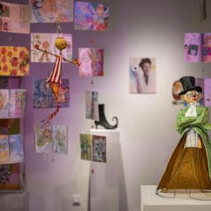 Kuva galleriasta, jossa etualalla lierihattupäinen veistos ja taka-alalla on katosta roikkuvia värikkäitä teoksia. 