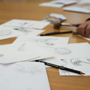 Drawing papers on the table. A hand holding a pencil.
