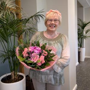 Vaaleahiuksinen hymyilevä Aija Karttunen seisoo viherkasvin vieressä. Hän pitelee käsissään suurta kukkakimppua, jossa on vaaleanpunaisia kukkia.
