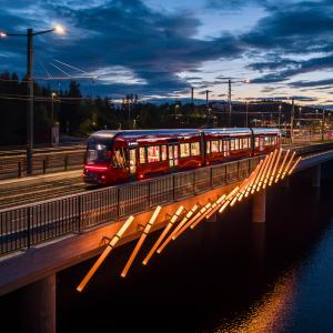 Kuvassa ratikka sillalla ja valoteos kyljessä, iltahämärässä.