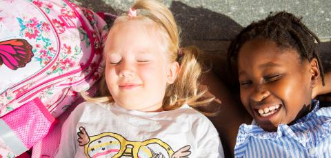 Kaksi lasta makaa maassa auringossa ulkona vierekkäin. Koulureppu on vieressä.