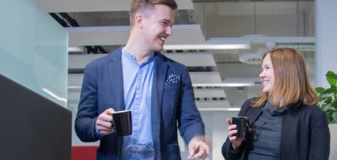 Avokonttorissa kävelee mies ja nainen kahvikupit käsissä. He katsovat toisiaan ja juttelevat.