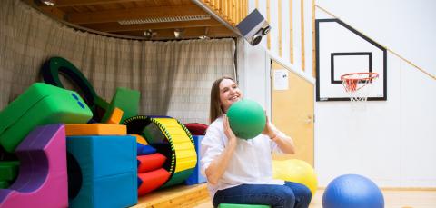 Nainen pitelee vihreää palloa käsissä liikuntasalissa. Taustalla leikkivälineitä.