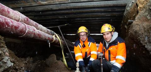 Kaksi asentajaa tunnelissa ja heidän vieressä on vesiputket. Miehillä on päällä työpuvut ja kypärät.