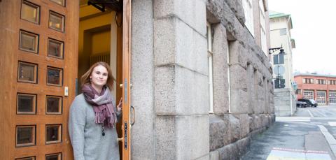 Nainen kurkkaa ulko-ovesta. Taustalla näkyy katua ja rakennuksen kulma.