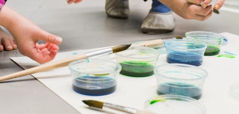 Sinistä ja vihreää vesiväriä läpinäkyvissä purkeissa. Lapsen kädet pitelevät sivellintä.