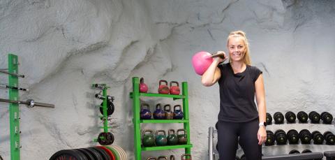 Henkilö pitelee isoa kahvakuulaa yhdellä kädellä käsi koukussa luolamaisessa kuntosalissa.