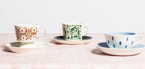 Kolme värikästä kahvikuppia on aseteltu vaaleanpunaiselle pellavapöytäliinalle.