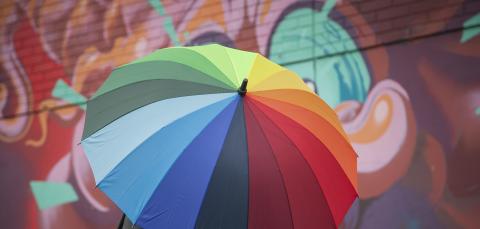 Ihminen seisoo monivärisen sateenvarjon alla suojassa.