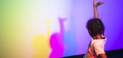 Lapsi käsi pystyssä, seinällä sateenkaaren värisiä varjoja.