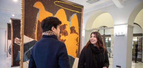 Suuressa galleriatilassa roikkuu lattiasta kattoon ulottovia ryijyä. Niiden edessä kaksi henkilöä juttelevat keskenään, toinen on katsojaan selin, toinen hymyilee ystävälleen.