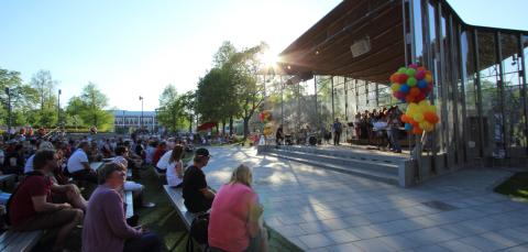 Aurinko paistaa kesäisessä maisemassa. Suurella ulkoilmalavalla esiintyy orkesteri. Yleisö istuu penkeillä lavan edessä.