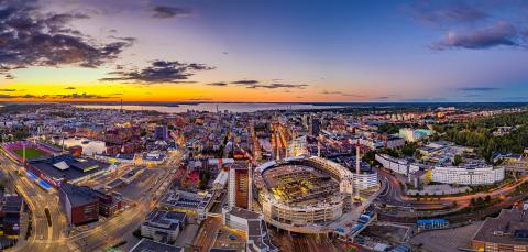 Ilmakuvaperspektiivi Tampereen keskustasta, missä näkyy rakennuksia ja viheralueita. Taustalla aurinko on laskemassa Näsijärven suuntaan.