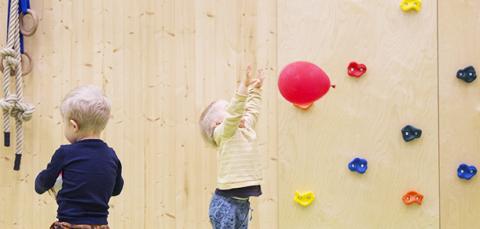Liikuntasalissa yksi aikuinen ja kolme lasta, joista yksi heittää ilmapalloa. Taustalla kiipeilyseinä.