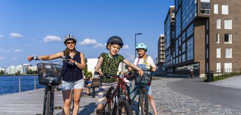 Aikuinen ja kaksi lasta pyöräilemässä kesäpäivänä Tampellan alueella Näsijärven rannassa. Kaikilla on pyöräilykypärä päässään.