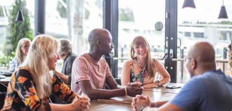 Neljä eri etnisistä taustoista tulevaa ihmistä keskustelevat saunaravintola Kuuman pöydän ääressä.