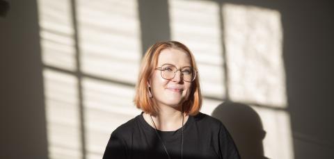 Hymyilevä henkilö auringonvalossa huoneessa, jonka seinälle henkilön taakse heijastuu ikkunakuvio valoin ja varjoin.