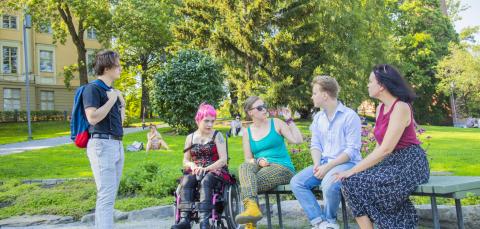 Joukko nuoria tamperelaisia aikuisia keskustelemassa ulkona Koskipuistossa kesällä. Osa heistä istuu, yksi seisoo ja yksi on pyörätuolissa.