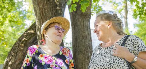 Kaksi  henkilöä seisoo lehdessä olevan puun alla ja katsoo toisiaan.