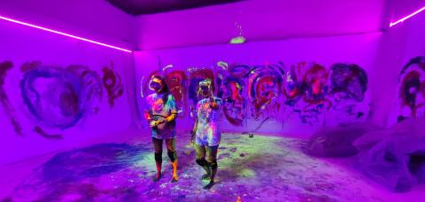 Kaksi maalinpeittämää esiintyjää keskellä violettia maalilla roiskittua tilaa.