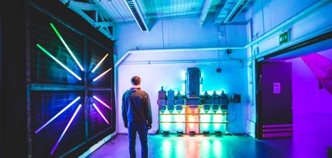 Takaapäin kuvattu mies on katselemassa valotaideteosta, jossa metallinen laite on valaistu vihreillä ja oranssilla valolla. Vasemmalla metalliverkkoseinällä on eri värisiä loisteputkia säteiksi aseteltuna. Huone on täynnä violettia, vihreää ja sinertävää valoa.