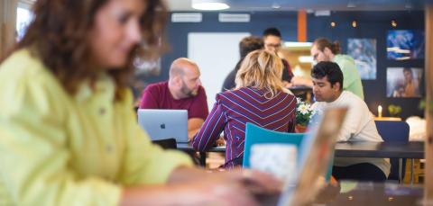 Henkilöitä työskentelemässä kahvilatilassa. Etualalla henkilö tietokoneella.