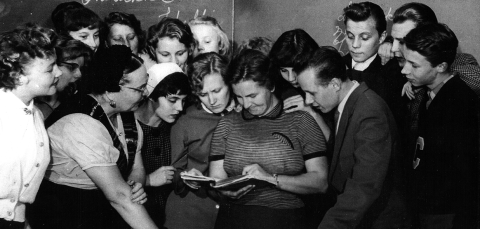 Joukko opiskelijoita on kokoontunut opettajan ympärille tutkimaan kirjaa.