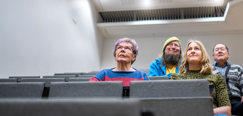 Neljä henkilöä istuu auditoriossa vierekkäin.