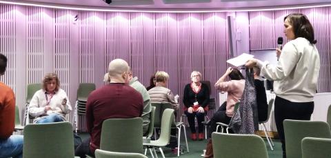 Aluetoimijaverkoston tapaamisessa tummahiuksinen nainen on pitämässä tervetulopuhetta mikrofoni kädessään. Kuulijat istuvat kehässä tuoleilla.
