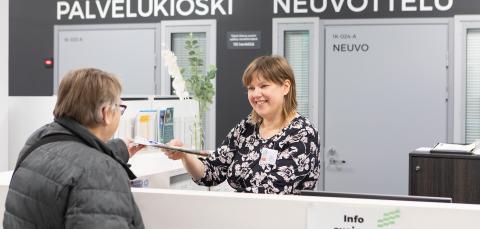 Tesoman hyvinvointikeskuksen infopisteellä hymyilevä työntekijä ojentaa esitettä asiakkaalle.