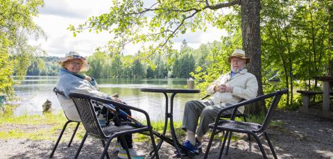 Kaksi ikääntynyttä henkilöä istuu puutarhatuoleilla pöydän ääressä järven rannalla.