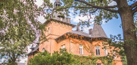 Tiilinen kartanorakennus alaviistosta kuvattuna kesäisessä maisemassa, edessä vihreää kasvillisuutta.