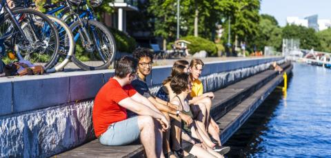 Joukko nuoria aikuisia istuu Tammerkosken Ratinanvuolteen reunalla kesäpäivänä. Heidän takanaan näkyy polkupyöriä.