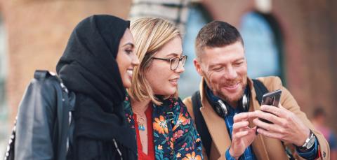 Kolme hymyilevää ihmistä istuu rinnakkain. Yksi heistä näyttää muille jotakin puhelimen näytöltä.