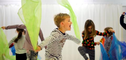 Lapset tanssivat värikkäiden huivien kanssa valkoisessa tilassa.